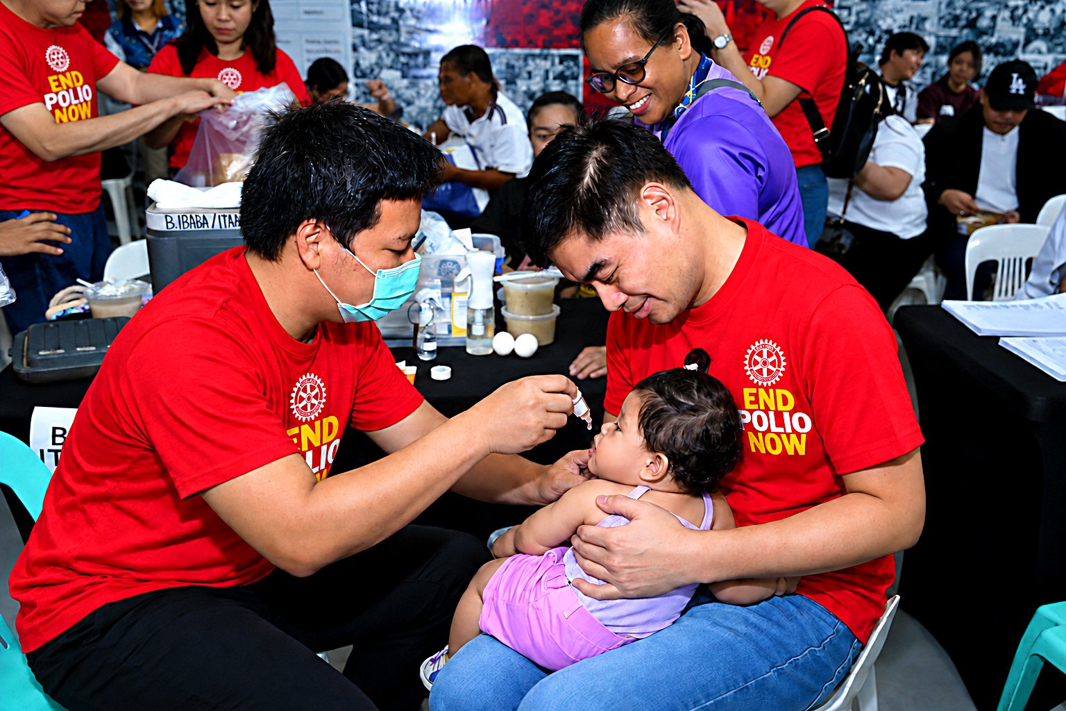 President Reggie Yu, in an End Polio Now shirt, holds a baby on his lap as a volunteer doctor gives her oral polio vaccine drops.